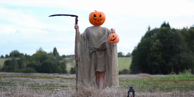 scarecrow with pumpkin head holding a sickle and a jack-o-lantern in a field with smiling face