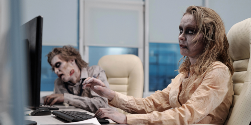 Two zombies sitting in arm chairs at a desk looking at a computer monitor and typing on their keyboards