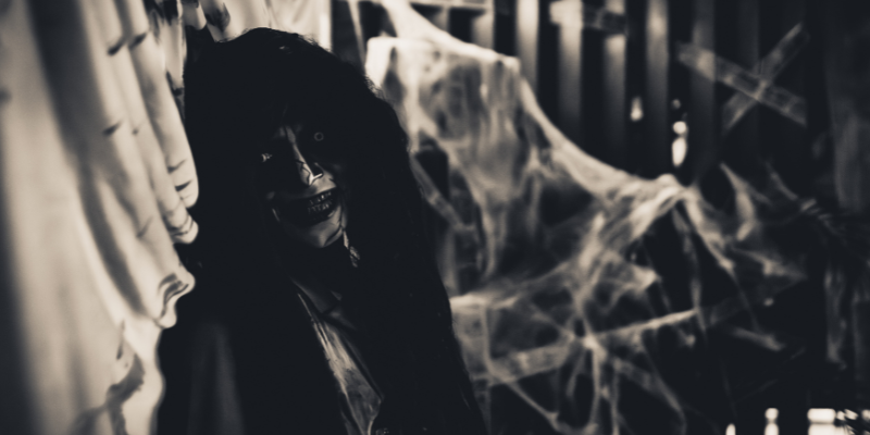 Black and white image of a scary woman cloaked in black in a cobweb filled room