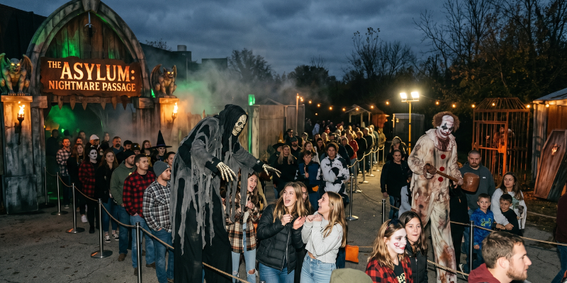 Nighttime scene outside a haunted house with a massive line of guests waiting to enter while scare actors interact with visitors in the queue
